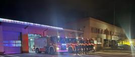 Strażacy Jednostki Ratowniczo-Gaśniczej numer 2 w Częstochowie stojąc w szeregu oddają hołd strażakowi, który zginął na służbie, w tle samochody strażackie z włączonymi niebieskimi sygnałami świetlnymi oraz budynek Jednostki Ratowniczo-Gaśniczej.
