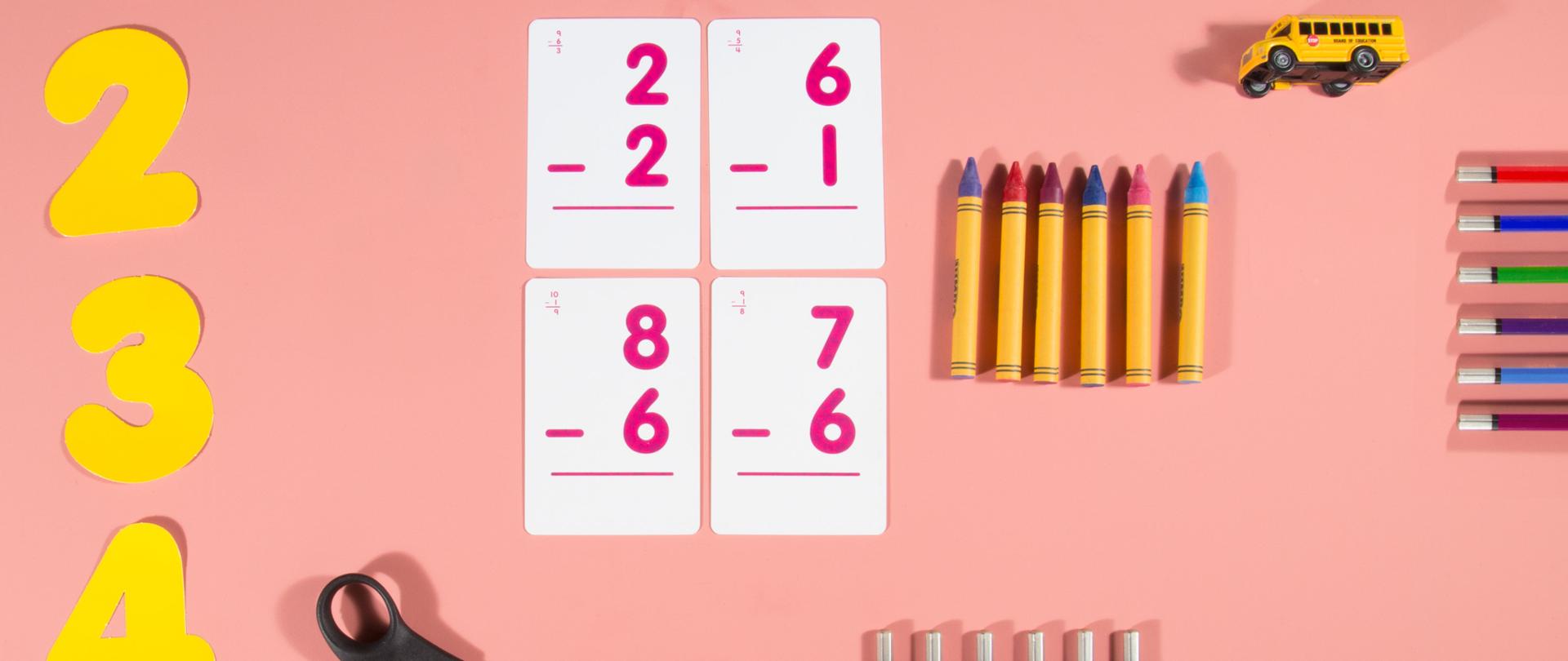 Na różowym tle wycięte z żółtej kartki cyfry, nożyczki, kredki, zabawkowy autobus i kolorowe kartki