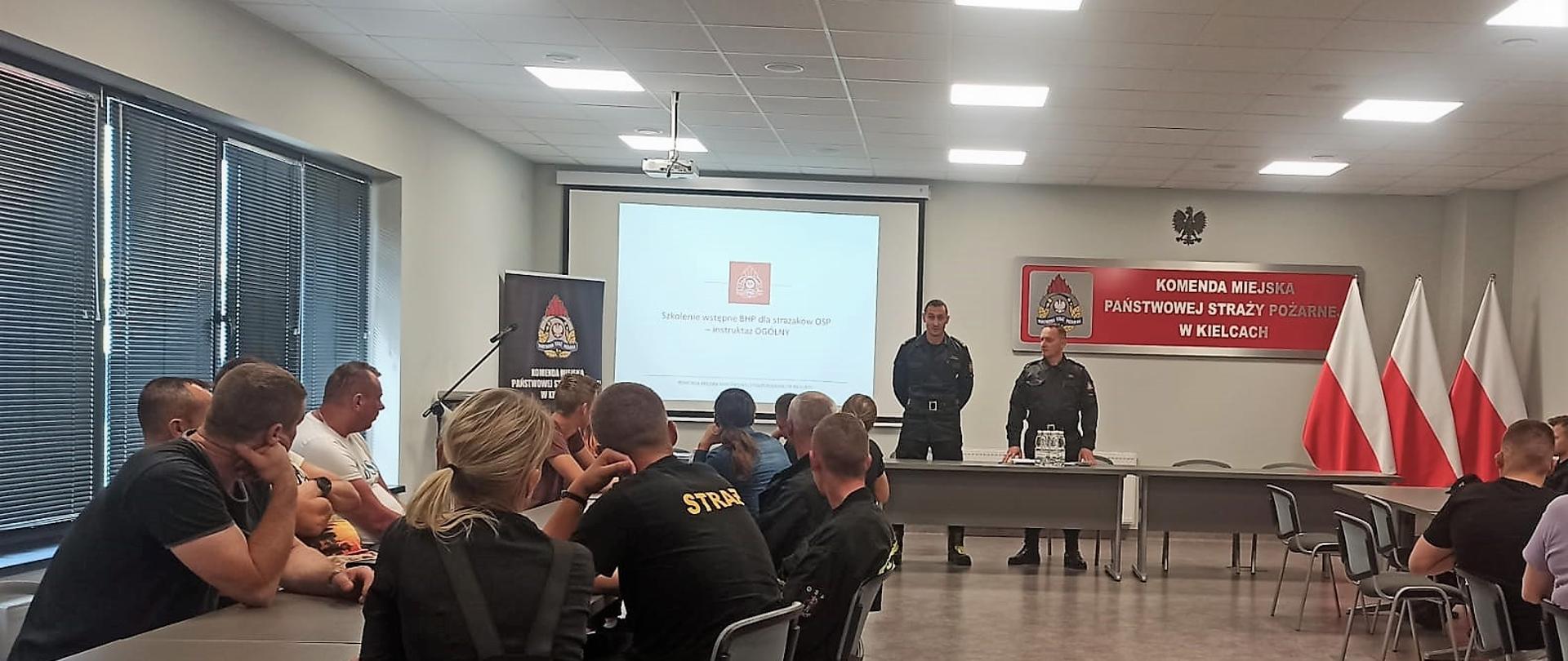 Na zdjęciu przedstawiono salę konferencyjną gdzie dobyło się pierwsze spotkanie uczestników kursu podstawowego dla strażaków ochotników. Stojący przy stole głównym Zastępca Komendanta Miejskiego Państwowej Straży Pożarnej w Kielcach mł. bryg. Michał Jankowski mówi do zebranych, a obok stoi funkcjonariusz z Wydziału Operacyjnego mł. kpt. Adam Kotaś.