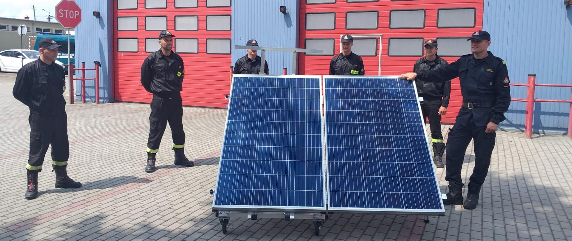 Zdjęcie przedstawia trenażer instalacji fotowoltaicznej oraz kilku strażaków w umundurowaniu koszarowym. Zdjęcie zrobione przed garażami JRG Jędrzejów, w tle bramy garażowe.