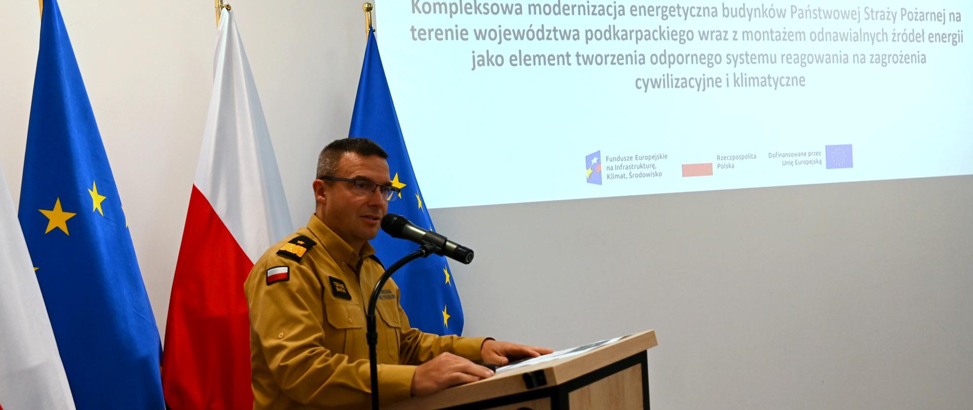 Nadbrygadier Tomasz Baran, komendant wojewódzki PSP, przemawia do mikrofonu przy mównicy. Ma na sobie piaskowy mundury służbowy. W tle widoczne flagi Polski i Unii Europejskiej oraz fragment ekranu z tytułem projektu dotyczącego modernizacji energetycznej budynków straży pożarnej na Podkarpaciu.