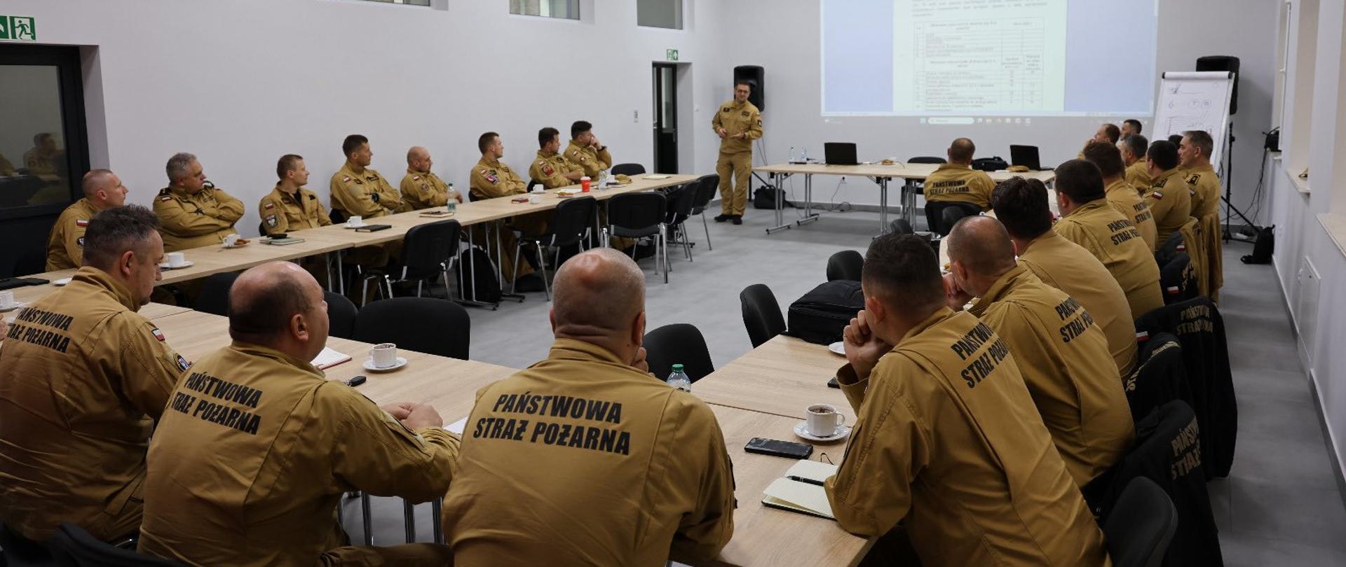 Grupa strażaków siedzi przy stole w sali konferencyjnej podczas odprawy dowódców jednostek PSP w Zachodniopomorskiem. Wszyscy ubrani są w piaskowe mundury. Sala jest jasno szara a sufit biały.