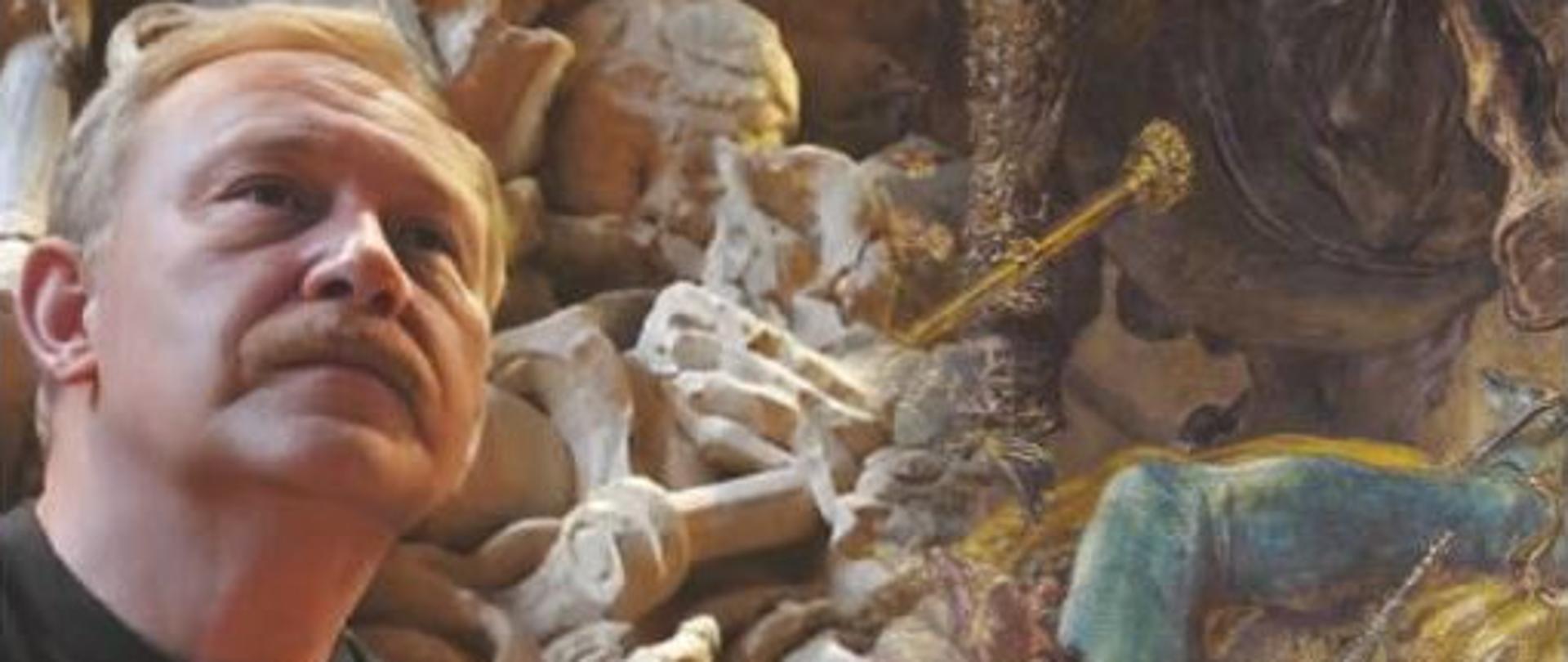 Na zdjęciu mężczyzna w ujęciu popiersiowym w układzie trzy czwarte na tle płaskorzeźby w surowym bezbarwnym drewnie odwzorowującej fragment obrazu Bitwa pod Grunwaldem