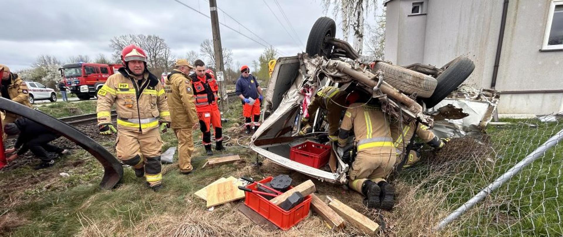 Centralny punkt zdjęcia stanowi rozbity biały bus leżący na dachu. Bus jest zdeformowany, pogięty. Przy aucie służby ratunkowe, strażacy i ratownicy medyczni. Wszyscy próbują wydobyć poszkodowanego.