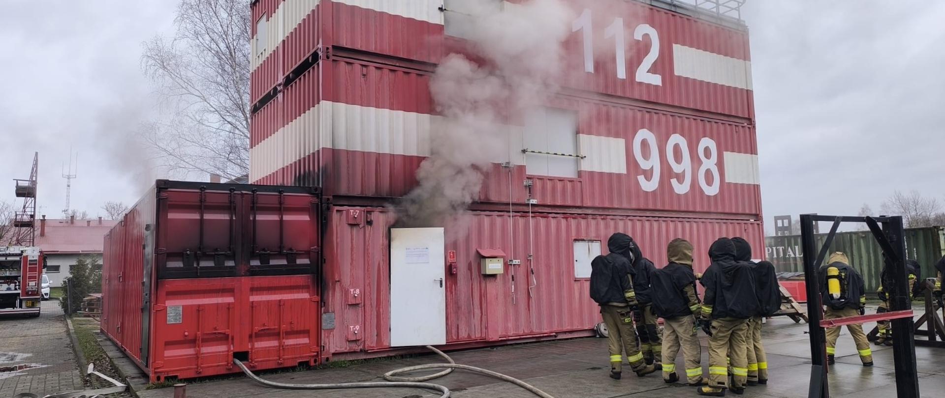 Widok ogólny trenażera ratwoniczego – z wnętrza wydobywa się dym, a grupa strażaków przygotowuje się do wejścia i prowadzenia działań gaśniczych.