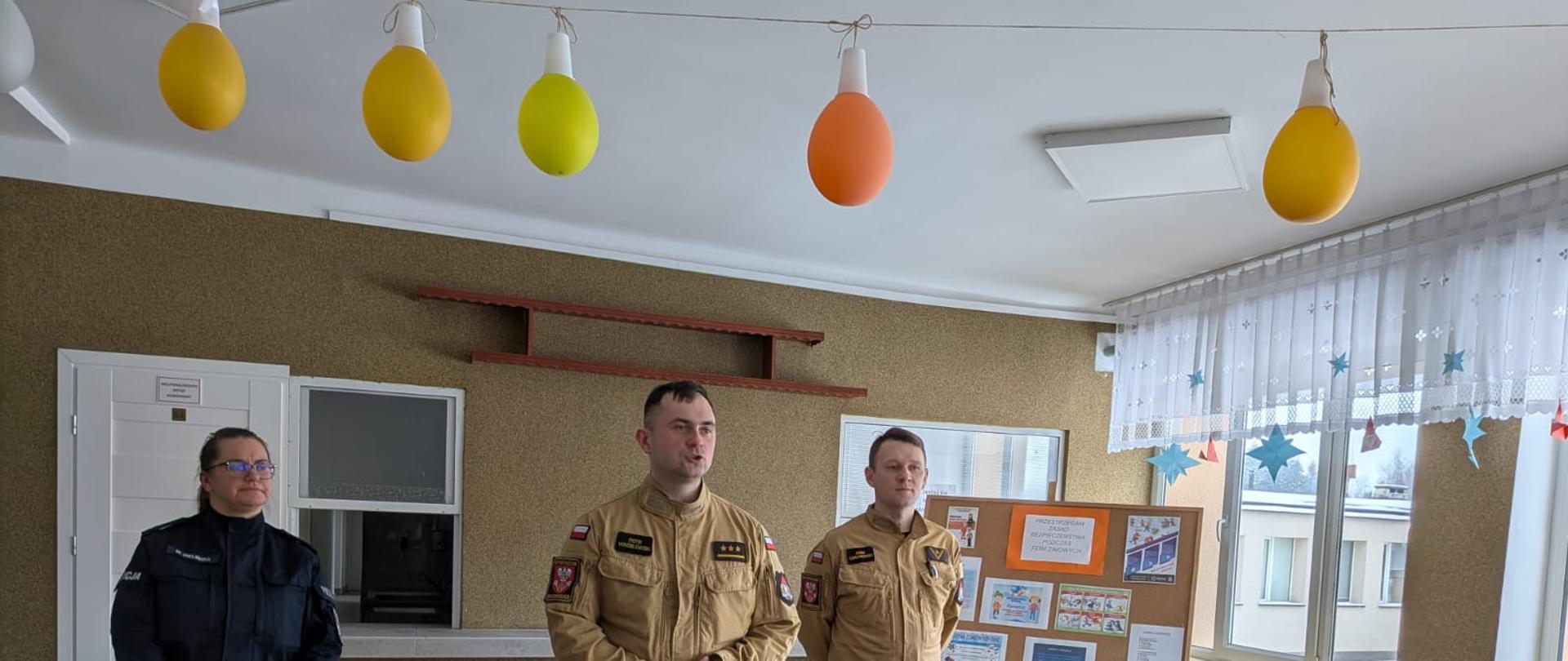 trzy osoby w mundurach służbowych stojące przodem do obiektywu, a za nimi, na krzesłach, siedzi grupa dzieci. Dwie osoby po prawej stronie to strażacy w piaskowym umundurowaniu koszarowym, z widocznymi dystynkcjami oraz emblematami na ramionach. Kobieta po lewej stronie ubrana jest w ciemnogranatowy lub czarny mundur policyjny. W tle widoczna jest tablica informacyjna, okna z zasłonami oraz dekoracje w postaci kolorowych, podłużnych balonów wiszących u sufitu.