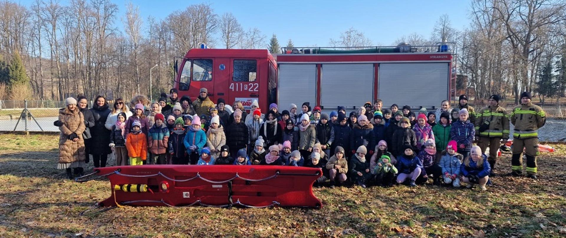 Kolorowa fotografia wykonana w pogodny dzień. Przedstawia grupę dzieci które uczestniczyły w pogadance i oglądały metody ratowania osób pod którymi załamał się lód. Obok dzieci strażacy w umundurowaniu specjalnym. W tle samochód pożarniczy.