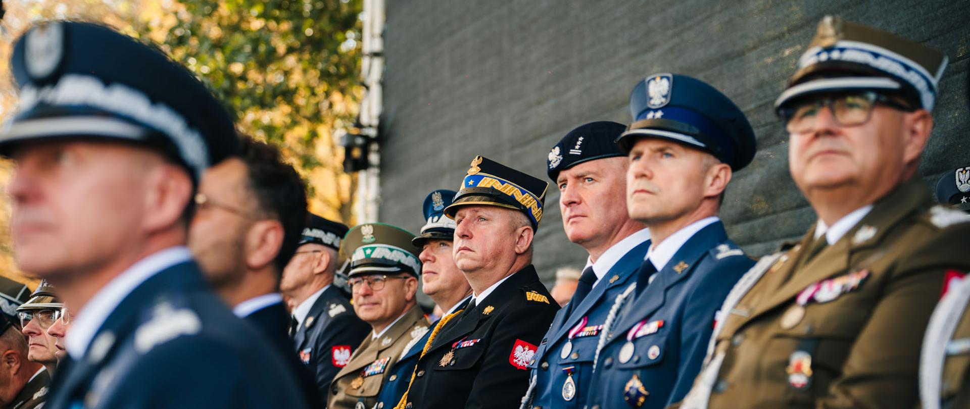 Przedstawiciele służb mundurowych siedzą na trybunie podczas oficjalnej uroczystości. W centrum widoczni są funkcjonariusze i oficerowie w galowych mundurach różnych formacji, m.in. wojska, policji i straży pożarnej.
