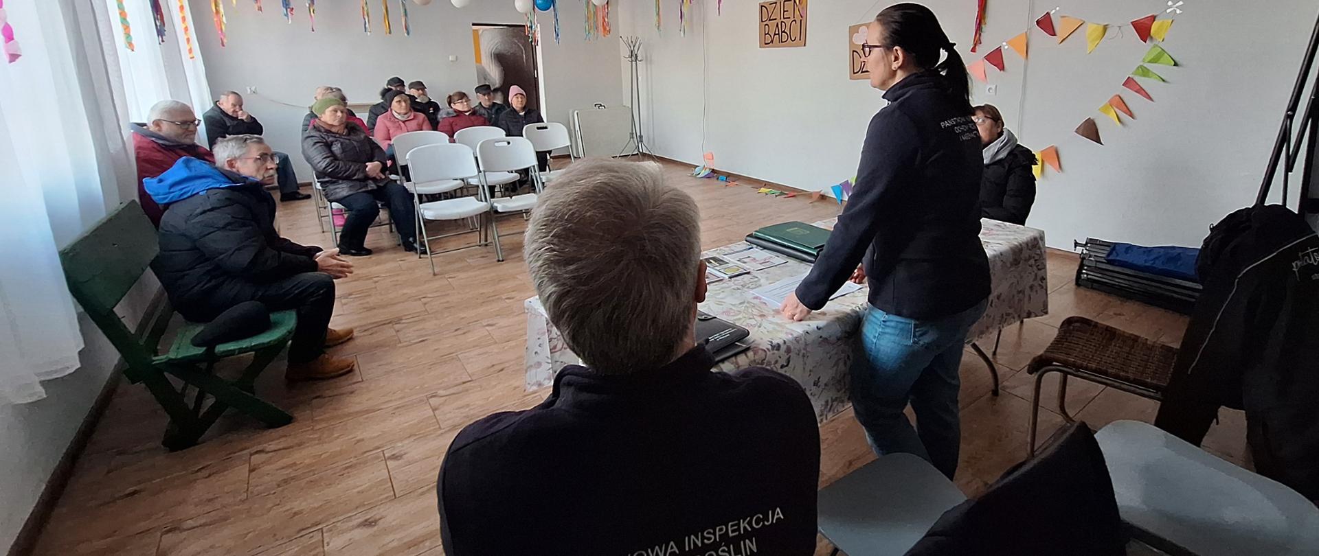 Od prawej przy stole siedzi jedna osoba, a druga stoi i przmawia sdo uczestników siedzących naprzeciw w kilku rzędach krzeseł