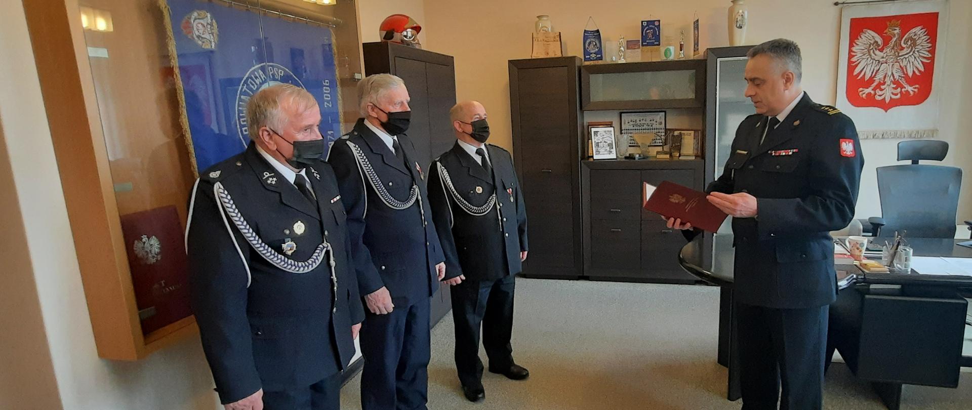 Gabinet komendanta powiatowego PSP. W tle widoczne godło i sztandar komendy. 3 druhów z OSP Wola Prażmowska w mundurach wyjściowych stoi przed Kondominatem Powiatowym PSP w Piasecznie. Komendant odczytuje treść decyzji o przyznaniu świadczenia.. 