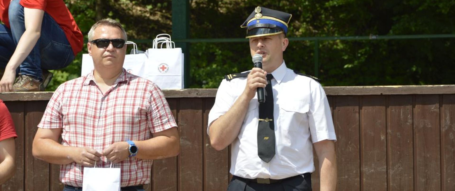 Na zdjęciu widać komendanta powiatowego oraz zastępce burmistrza w Ustrzykach Dolnych. Komendant ubrany jest w białą koszulę, krawat, czarne spodnie oraz czapkę rogatywkę. Zastępca burmistrza natomiast jest w koszuli w kratę oraz spodnie jeansowe i ciemne okulary. Komendant trzyma w ręku mikrofon i przemawia. Za nimi znajduje się scena "Parku pod Dębami"