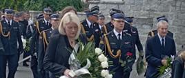 Uczestnicy pogrzebu w przeważającej większości w mundurach strażackich idą w kondukcie pogrzebowym. W tle zabudowania miejskie.