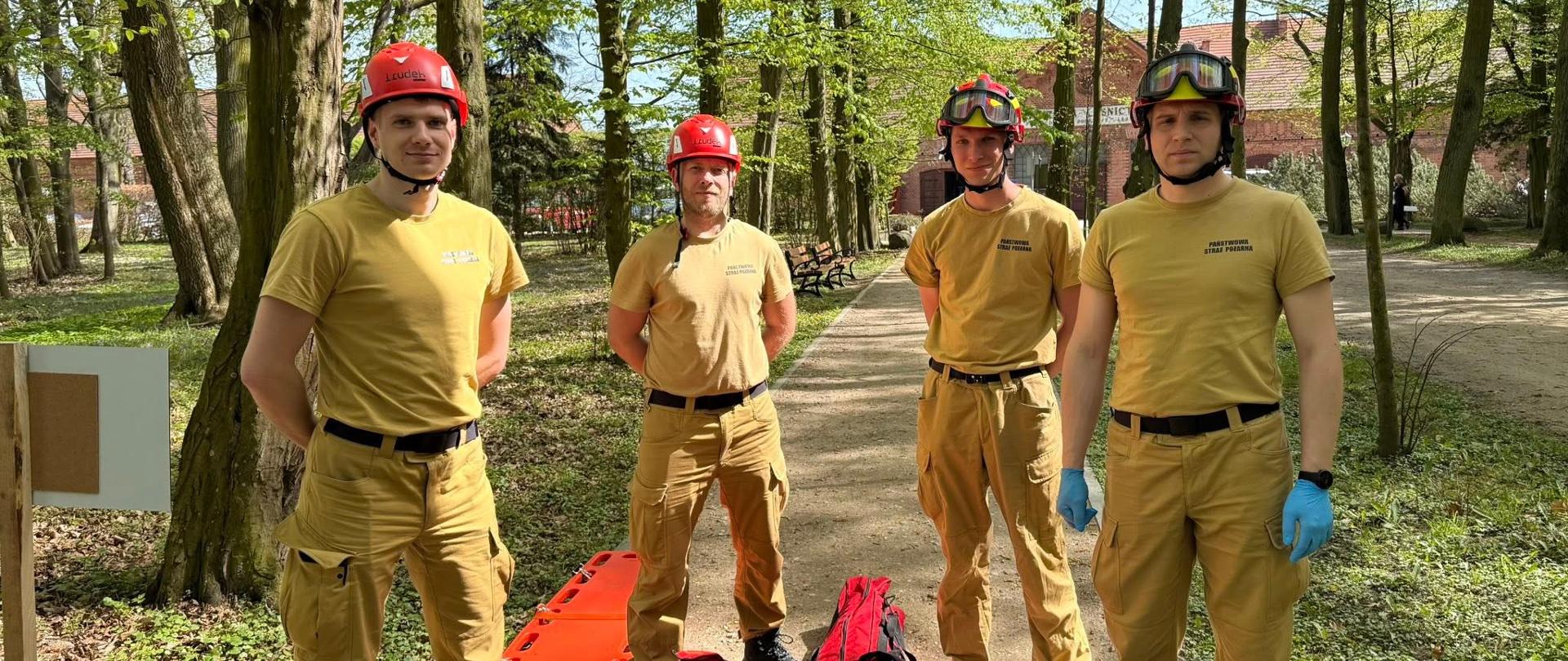 Na zdjęciu widzimy czterech mężczyzn ubranych w jednolite mundury w kolorze piaskowym — mają na sobie koszulki, bojówki oraz czarne buty. Na głowach czerwone hełmy ochronne z goglami. Jeden z nich (po prawej stronie) ma na rękach niebieskie rękawiczki ochronne.
Stoją na leśnej ścieżce wśród zielonych drzew — widać, że jest wiosna lub wczesne lato. Na ziemi przed nimi leżą czerwone torby ratunkowe i pomarańczowa deska ortopedyczna, używana zwykle do transportu rannych. W tle widać parkowe ławki i budynek z czerwonej cegły. Wygląda na to, że są to ratownicy lub uczestnicy szkolenia z zakresu ratownictwa.