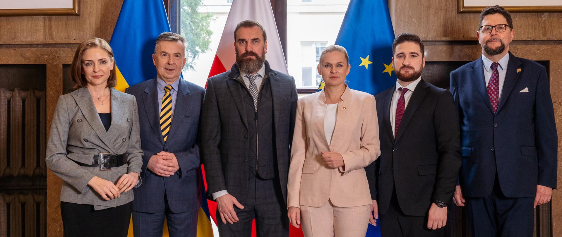Zdjęcie zbiorowe, na tle okna stoją i flag Polski, Ukrainy i UE stoi minister Nowacka, minister Wieczorek i wiceminister Mucha oraz trzech mężczyzn w garniturach