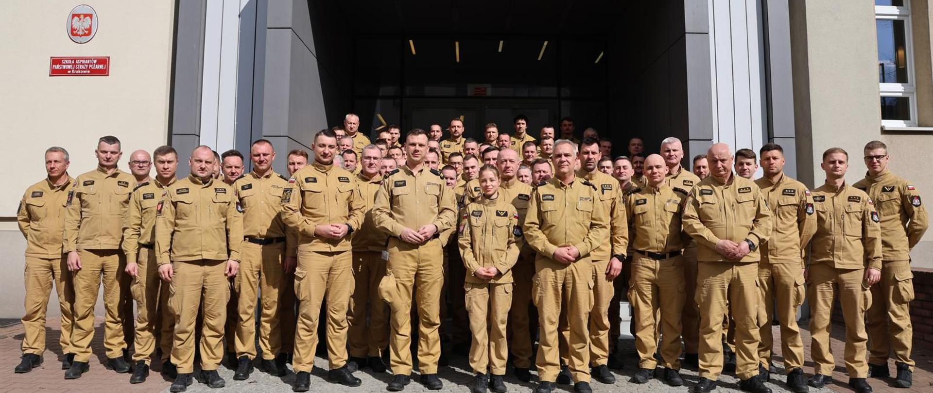 Uczestnicy warsztatów przed budynkiem Szkoły.
Pamiątkowe zdjęcie grupowe kilkudziesięciu strażaków w piaskowych mundurach służbowych. Stoją przed wejściem głównym do Szkoły Aspirantów PSP w Krakowie. Na elewacji widoczne godło Polski i tablica informacyjna szkoły.
