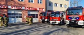 Strażacy stoją naprzeciw siebie w dwuszeregu po każdej stronie na placu przed garażami. Po prawej stronie ustawione są samochody strażackie