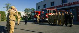 Strażacy w pododdziałach oddają honor podczas podniesienia flagi na maszt, ustawieni na tle budynku komendy oraz samochodów pożarniczych na przeciwko siebie
