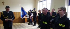 Widok z przodu. Po lewej stronie na pierwszym planie komendant powiatowy PSP odczytuje tekst ślubowania. Po prawej stronie dwoje strażaków z uniesionymi rękoma powtarza tekst ślubowania. Pośrodku na drugim planie widoczny poczet sztandarowy JRG.