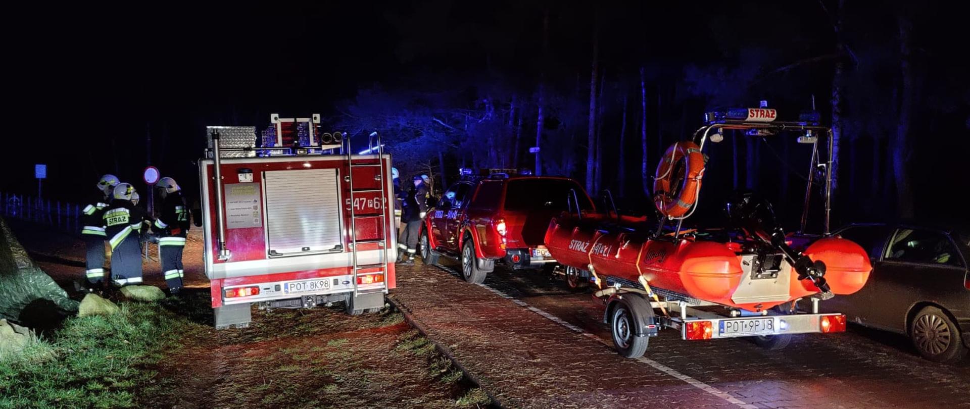 Zdjęcie przedstawia służby ratownicze , które biorą udział w poszukiwaniach osoby zaginionej na zalewie "Blewązka" w Kobylej Górze.
Na pierwszym planie widać łódź ratowniczą na przyczepce a obok pojazd ratowniczy przy którym znajdują się strażacy w ubraniach specjalnych. Zdjęcie wykonane w godzinach nocnych przy utwardzonym dojeździe do zalewu. 