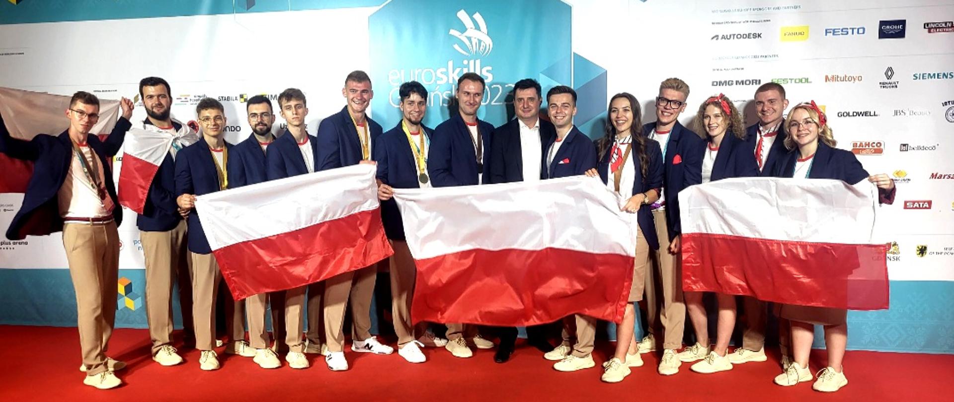 Na tle niebieskiej ściany stoi grupa kilkunastu osób w granatowych marynarkach, trzymają przed sobą trzy rozpostarte polskie flagi.