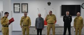 Strażak w umundurowaniu koloru musztardowego trzyma czerwoną teczkę i czyta przed nim stoi kierownictwo Komendy Wojewódzkiej Państwowej Straży Pożarnej w Gdańsku oraz mężczyzna w garniturze.