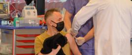 Na zdjęciu widzimy mężczyznę który siedzi na krzesełku i przygotowuje się do szczepienia. Mężczyzna ubrany jest w piaskowy mundur , rękaw lewej ręki zawinięty do góry, czarna maseczka na twarzy. Przy mężczyźnie stoi lekarz ubrany w biały kitel, czarne spodnie, brązowe buty, biała maseczka na twarzy. Lekarzy trzyma strzykawkę i dokonuje szczepienia. Obok lekarza stoi mężczyzna z pomocy medycznej ubrany w niebieski kitel, biała maseczka czarne buty z biała podeszwą, który asystuje lekarza. Całość dzieje się w pomieszczeniu, dalej w kadrze widzimy okna od podłogi do sufitu za oknami widzimy biało-czerwone krzesełka stadionowe. Za mężczyznami stoi szafka na kółkach koloru szarego. Podłoga w pomieszczeniu koloru brązowego. 