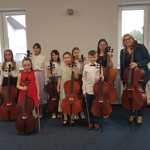 Zdjęcie przedstawia uczniów klasy wiolonczeli Pani Urszuli Właźniak. Od prawej: Kinga Halecka, Hanna Kowalewska, Weronika Krzyżewska, Sonia Lubartowicz, Hanna Dobrzycka, Nikola Matysiewicz, Adam Rogowski, Aleksandra Jasińska, p. Urszula Właźniak. Wszyscy trzymają w dłoniach wiolonczele. Ubrani są w odświętne stroje. W tle okna na białej ścianie, dekoracyjne obudowy grzejników w kolorze białym oraz granatowa wykładzina. 