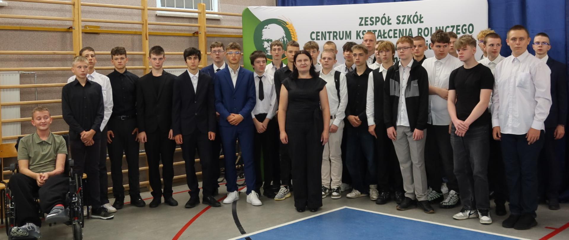 Grupa uczniów oraz nauczycielka pozują na tle baneru z nazwą i logotypem szkoły.