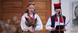 Dwoje młodych uczestników wydarzenia kulturalnego stoi przy mikrofonach i odczytuje tekst z przygotowanych teczek. Oboje są ubrani w tradycyjne stroje ludowe: dziewczyna ma kolorowy wianek, haftowany gorset i wzorzystą spódnicę, a chłopiec ciemny strój z czerwonymi ozdobami oraz tradycyjne nakrycie głowy. W tle znajdują się drewniane drzwi oraz dekoracje nawiązujące do wiejskich zbiorów, w tym słoneczniki, kosz z owocami i snopki siana