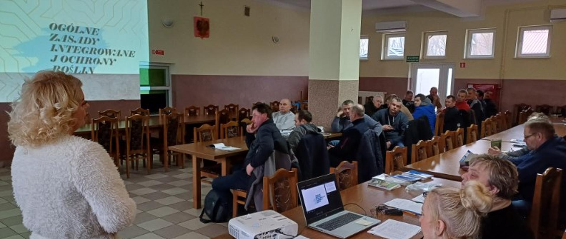 Kobieta ubrana w jasny strój stoi przed grupą ludzi siedzącą przy stolikach.
Po prawej stronie widać dwie kobiety siedzące przed laptopem. Obok laptopa leży projektor, który wyświetla prezentacje na ścianie. W pomieszczeniu znajdują się długie stoły i dużo krzeseł. Ściany są koloru brązowo-żółtego. W pomieszczeniu jest dużo małych okien.
