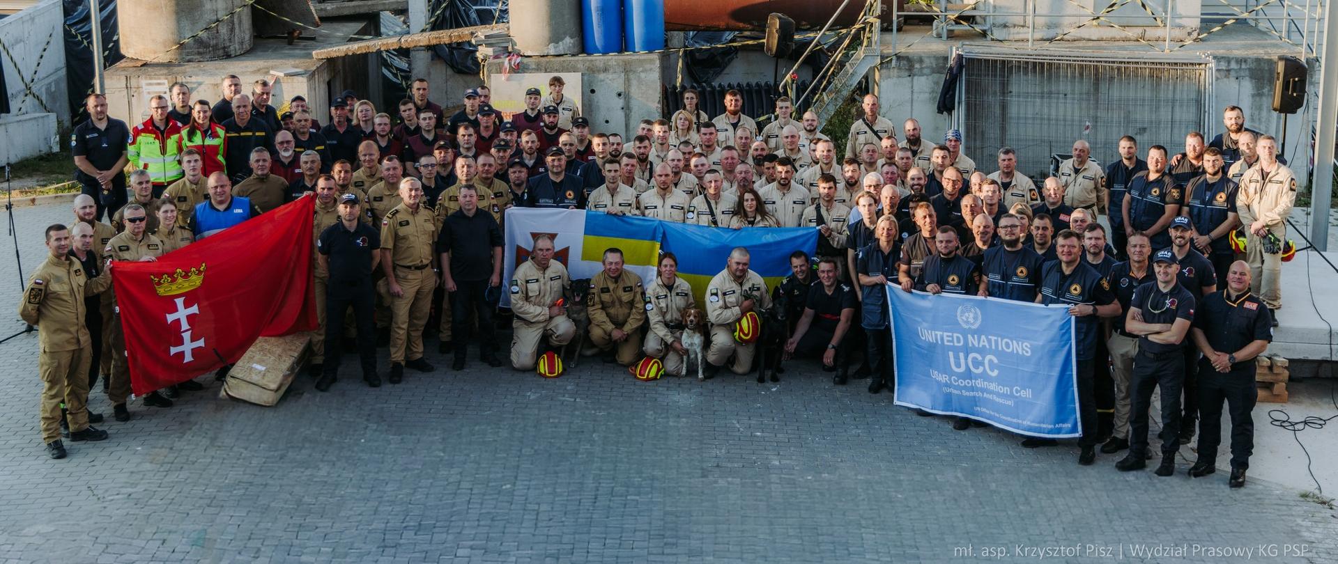 Strażacy pozują do wspólnego zdjęcia na tle gruzowiska.