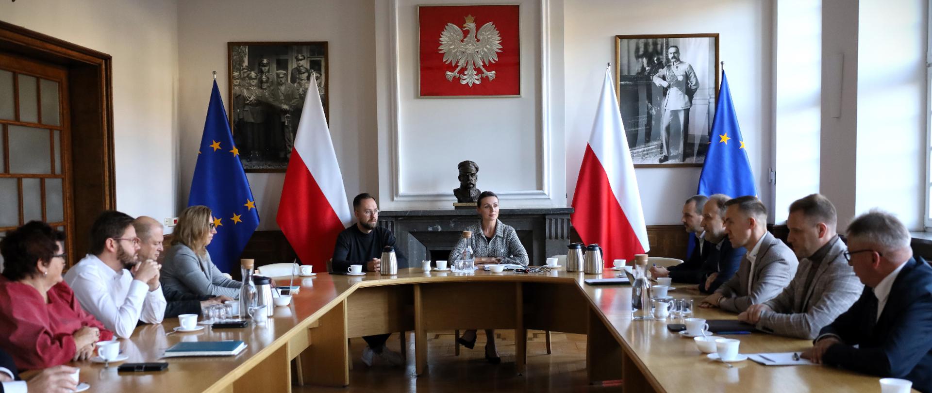 kilkanaście osób siedzi przy stole i rozmawia. Na ścianie godło Polski i portrety marszałka Piłsudskiego