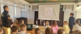 Spotkanie szkoleniowe na pierwszym planie prowadzona prezentacja. Po prawej stronie funkcjonariuszka policji po lewej funkcjonariusz straży pożarnej. Prezentacji przysłuchują się dzieci.