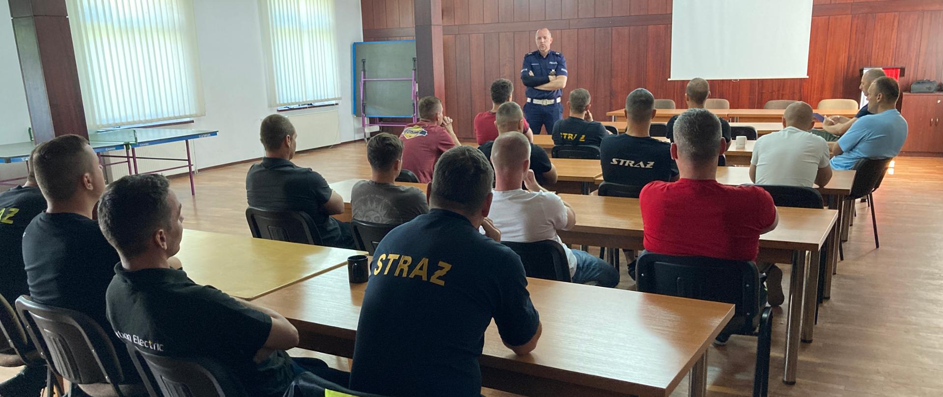 Naczelnik prowadzi szkolenie dla strażaków. Strażacy siedzą w sali szkoleniowej, natomiast naczelnik stoi. 