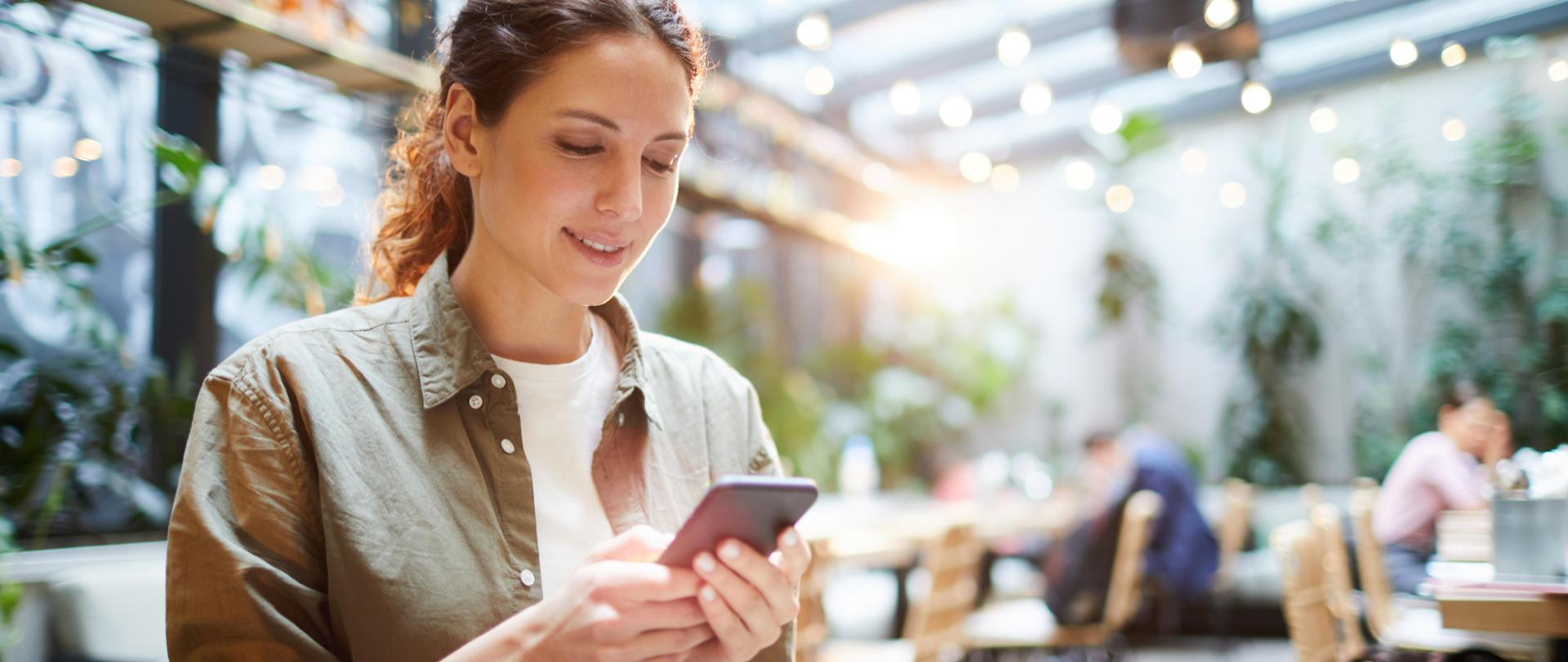 Kobieta z telefonem siedzi przy stoliku w kawiarni, na blacie kawa i talerz z ciastkiem.