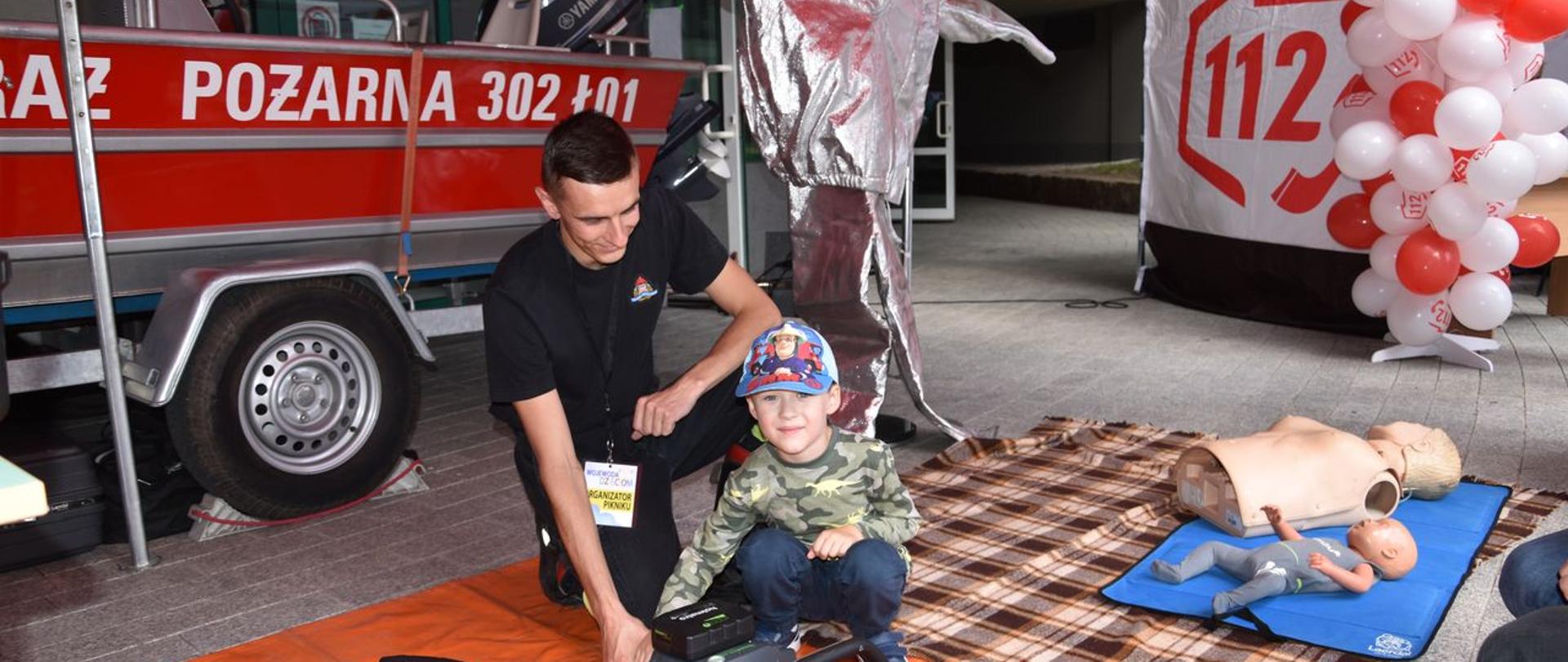 Na pierwszym palenie widzimy strażaka z dzieckiem – uczestnikiem pikniku, który bawi się narzędziami do uwalniania ludzi z pojazdów które uległy wypadkowi. Narzędzia leżą na pomarańczowej macie z napisem Holmatro. W tle strażacka łódź ratunkowa, ubranie żaroodporne, manekiny do nauki pierwszej pomocy oraz stoisko Centrum Powiadamiania ratunkowego.