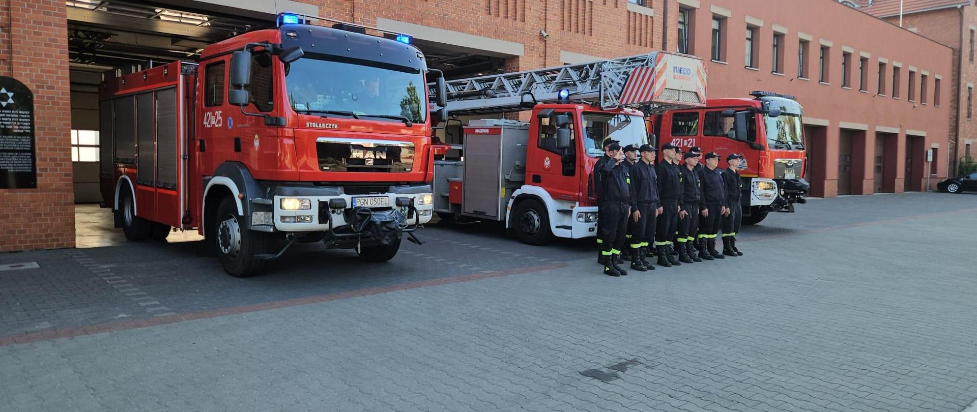 Strażacy w ciemnych mundurach stoją przed zaparkowanymi przed garażem komendy samochodami