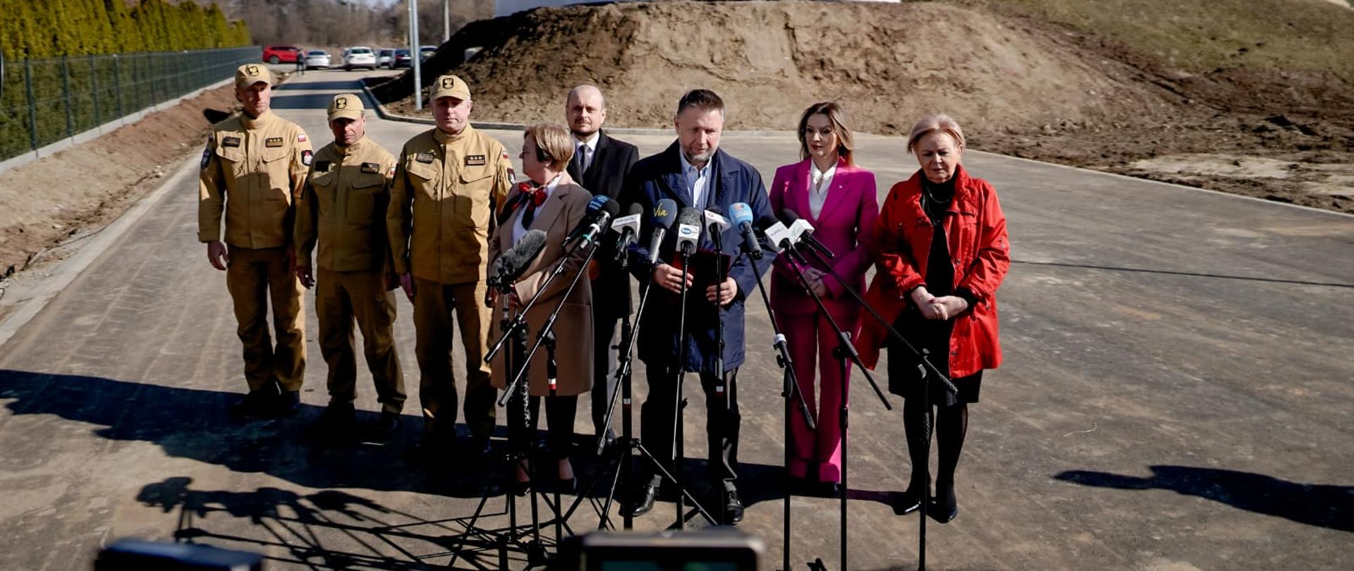 Konferencja prasowa przed dużymi zbiornikami wodnymi; grupa funkcjonariuszy Państwowej Straży Pożarnej i przedstawicieli władz stoi przed mikrofonami na otwartym terenie budowy.