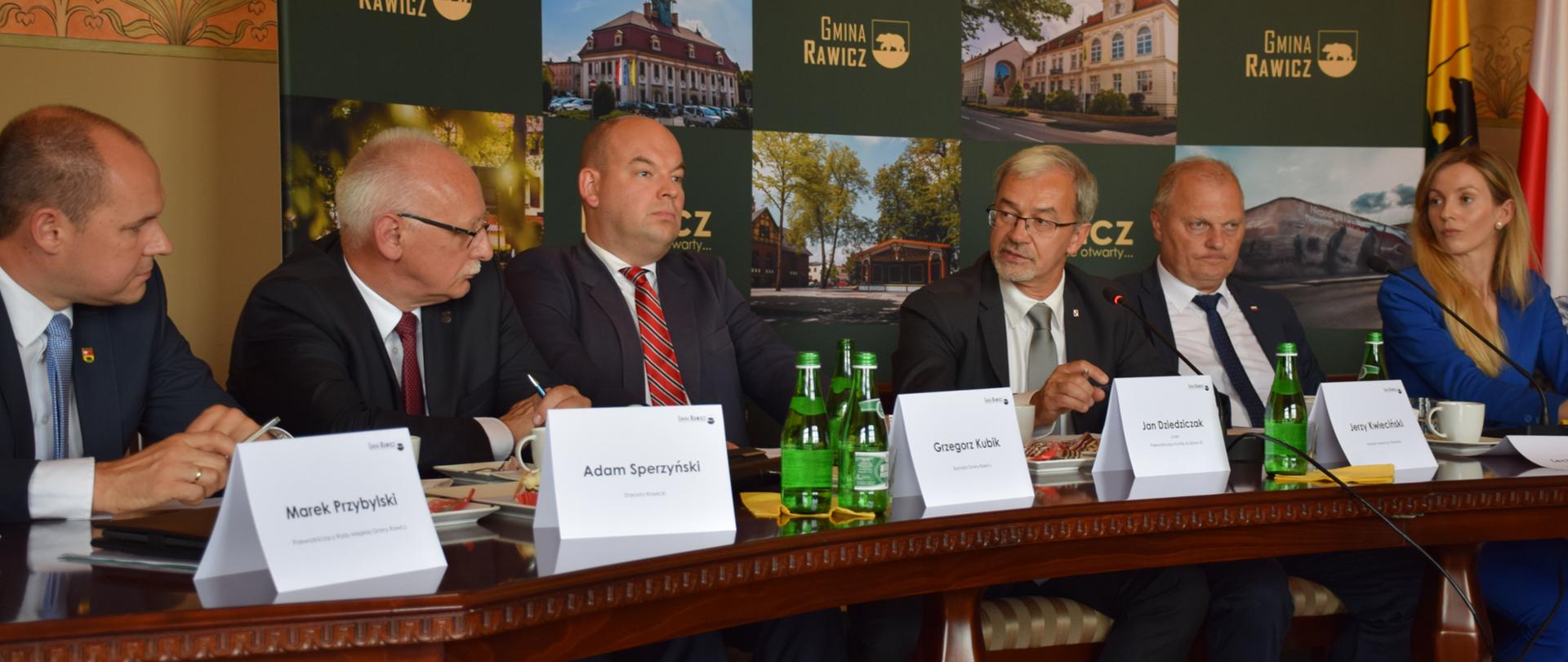 Sześć osób przy długim stole, z tyłu ścianka promocyjna gminy Rawicz, na stole butelki z wodą i koziołki z nazwiskami uczestników spotkania, trzeci od prawej minister Kwieciński