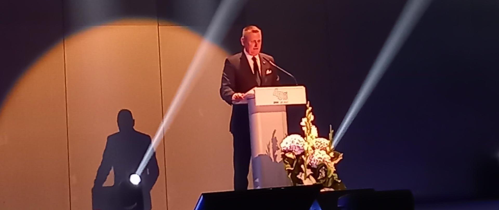 Na zdjęciu znajduje się ubrany w ciemny garnitur mężczyzna. Stoi on na scenie, przed mównicą i jest w trakcie wystąpienia publicznego. 