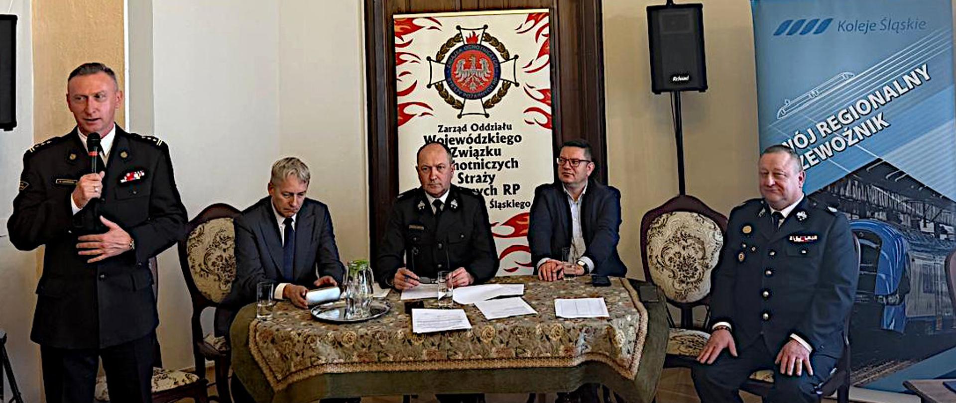 Zdjęcie przedstawia wnętrze sali konferencyjnej, gdzie odbywa się spotkanie. W centrum kadru znajduje się duży stół, nakryty jasnym obrusem w delikatny, kwiatowy wzór. Przy stole siedzi pięciu mężczyzn. Po lewej stronie kadru, nieco z przodu, stoi śląski komendant wojewódzki st. bryg. Damian Legierski, w ciemnym mundurze wyjściowym z dystynkcjami, trzyma mikrofon i przemawia. Obok niego, po lewej stronie stołu, siedzi mężczyzna w ciemnym garniturze i jasnej koszuli, z rozłożonymi dokumentami przed sobą. W centralnej części zdjęcia, za stołem, wisi duży, ozdobny sztandar z białym orłem na czerwonym tle. Poniżej orła widnieje napis "Zarząd Oddziału Wojewódzkiego Związku Ochrony Przeciwpożarowej Straży RP Śląskiego". Po obu stronach sztandaru stoją wysokie, wąskie banery reklamowe. Przy stole, po prawej stronie, siedzi dwóch mężczyzn w mundurach, podobnych do tego, który stoi z mikrofonem. Mężczyzna najbliżej środka kadru ma na sobie ciemny garnitur i okulary. Wszyscy mężczyźni siedzą prosto, z uwagą skierowaną na osobę mówiącą.