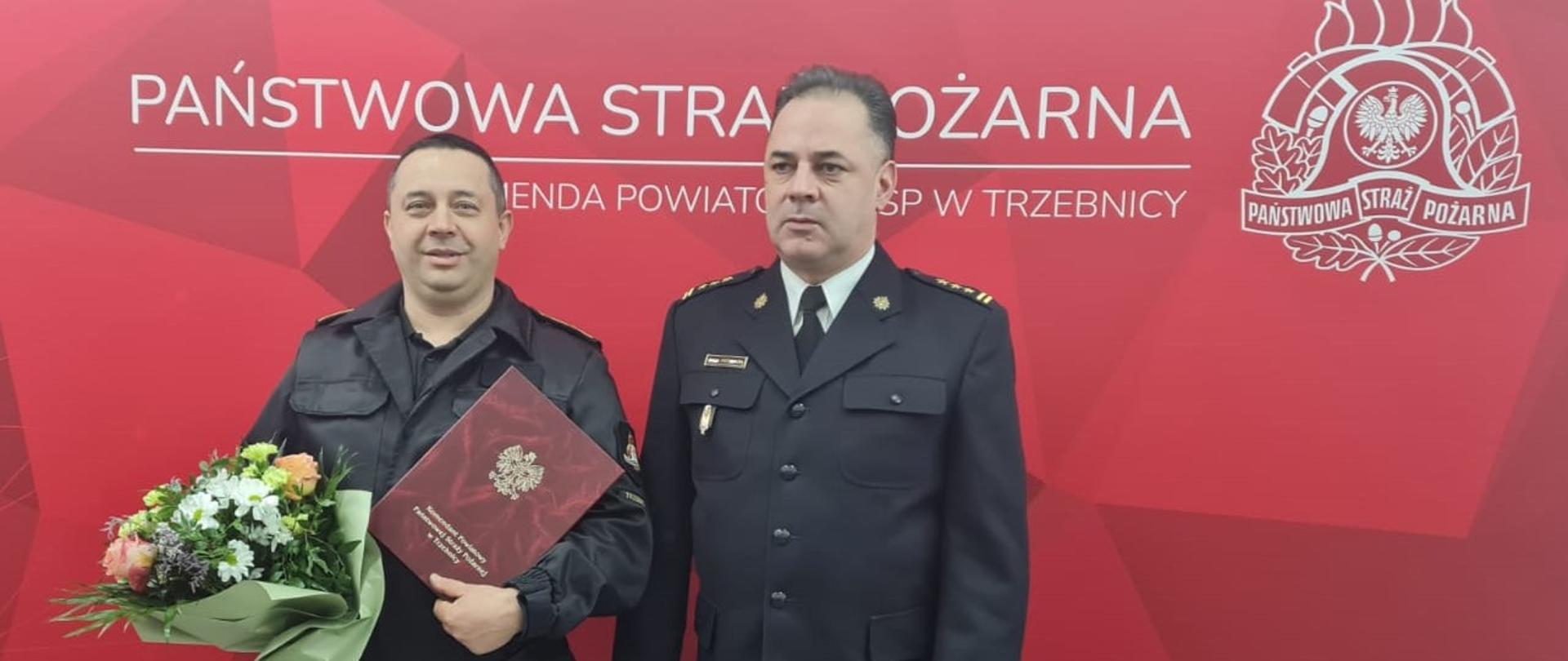Dwóch mężczyzn, na czerwonym tle, z logiem PSP. Komendant Powiatowy z odchodzącym na emeryturę strażakiem
