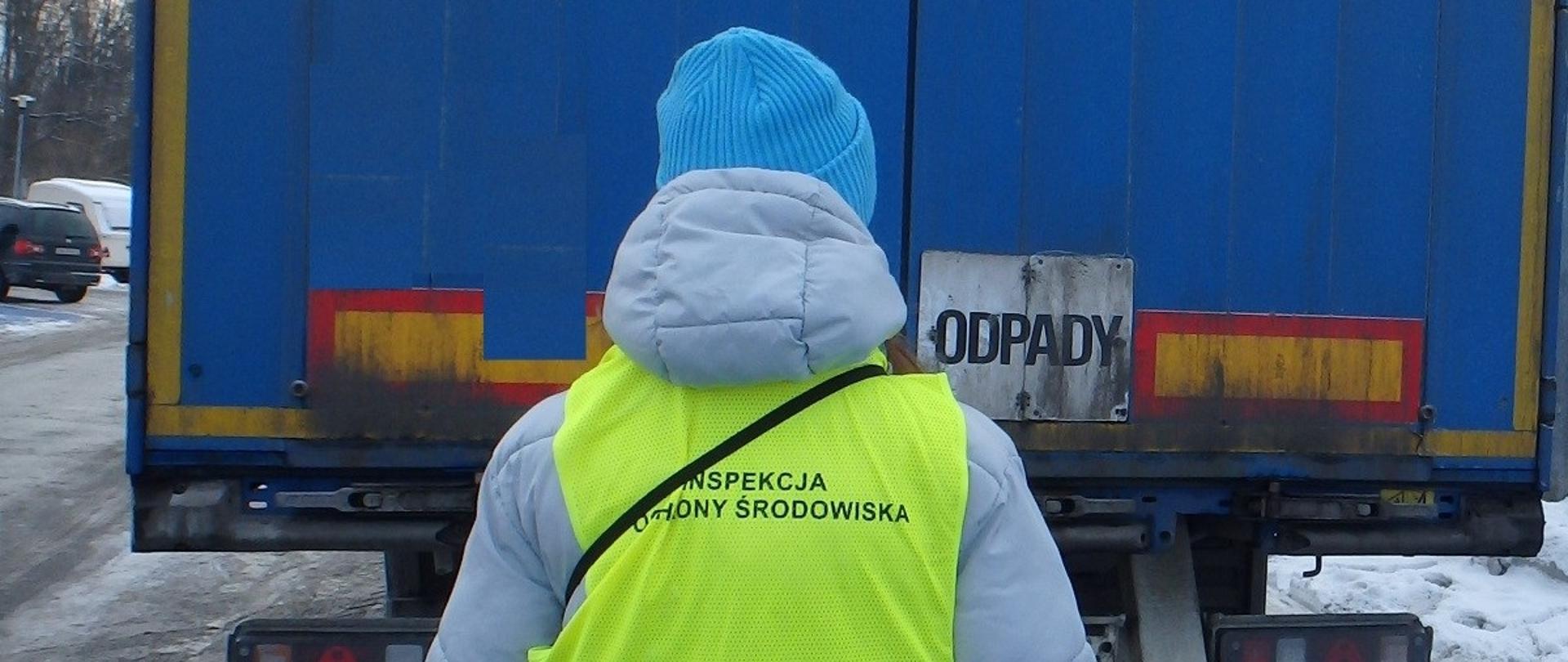 Osoba plecami do zdjęcia, ubrana w szarą, zimową kurtkę, niebieską czapkę, jeansy i z założoną kamizelką odblaskową z napisem Inspekcja Ochrony Środowiska. Przed osobą stoi zaparkowany samochód ciężarowy z naczepą w kolorze niebieskim i tabliczką z napisem odpady. W tle widać na ziemi śnieg i drugi zaparkowany samochód ciężarowy. 