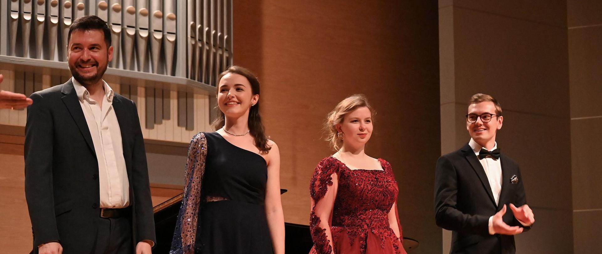 Na scenie sali koncertowej stoi czworo artystów. Od lewej: mężczyzna w ciemnym garniturze i białej koszuli (bez krawata), uśmiecha się; kobieta w granatowej sukni na jedno ramię, z rękawem z siateczki ozdobionej koralikami, uśmiecha się i patrzy w górę; kolejna kobieta w obszernej, bordowej sukni z bogatym zdobieniem i długimi koronkowymi rękawami (ta sama, co na poprzednim zdjęciu), patrzy przed siebie; i po prawej mężczyzna w czarnym smokingu z muszką i okularami, uśmiecha się i składa dłonie do oklasków. Z tyłu, po lewej, widać fragment dużego fortepianu oraz rzędy piszczałek organowych. Na pierwszym planie, u dołu, widoczny jest fragment dekoracji kwiatowej. W prawym dolnym rogu znajduje się znak wodny „POWIAT JAROSŁAWSKI”.