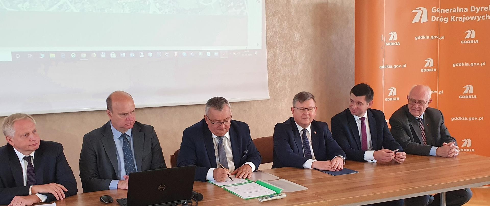 Na zdjęciu widać sześciu mężczyzn siedzących przy stole. Wszyscy panowie ubrani są w ciemne garnitury, mają przed sobą dokumenty.