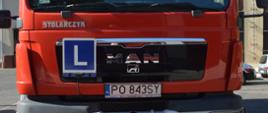 Szkolenie kierowców. Plac Komendy Powiatowej PSP w Rawiczu. Na tle budynku komendy stoi samochód kwatermistrzowski koloru czerwonego, który będzie wykorzystywany do zajęć praktycznych. Widoczny jest jego przód. Na kabinie, z lewej strony nazwy marki "MAN" umieszczona jest niebieska tabliczka z białą obwódką i białą literą "L" - oznaczającą samochód przeznaczony do nauki jazdy.