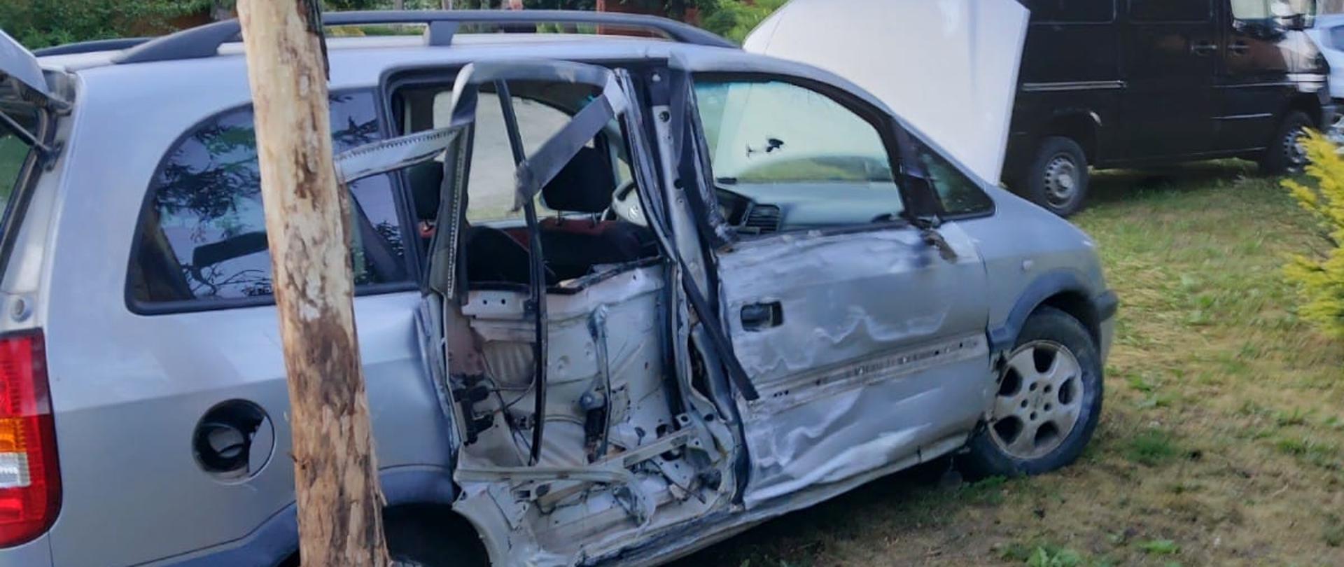 Na zdjęciu uszkodzony samochód osobowy koloru srebrnego obok drzewa za nim stoi inny samochód czarny bus