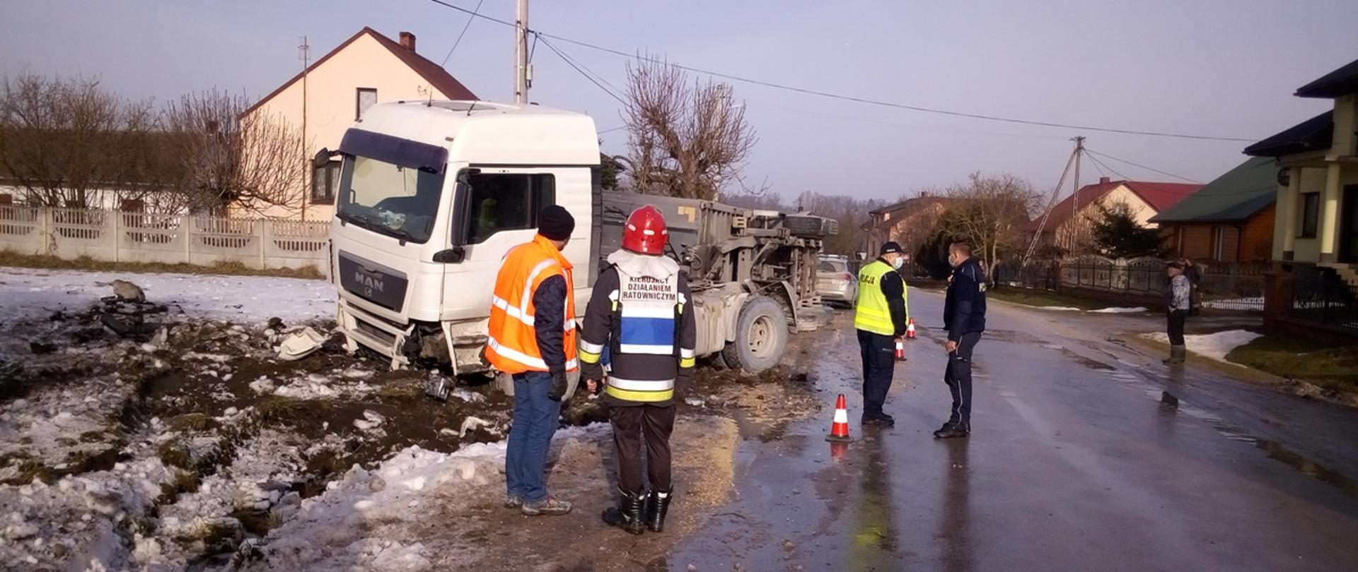Uszkodzony samochód ciężarowy stoi po lewej stronie na poboczy i w rowie, straż pożarna i policja prowadzą czynności.