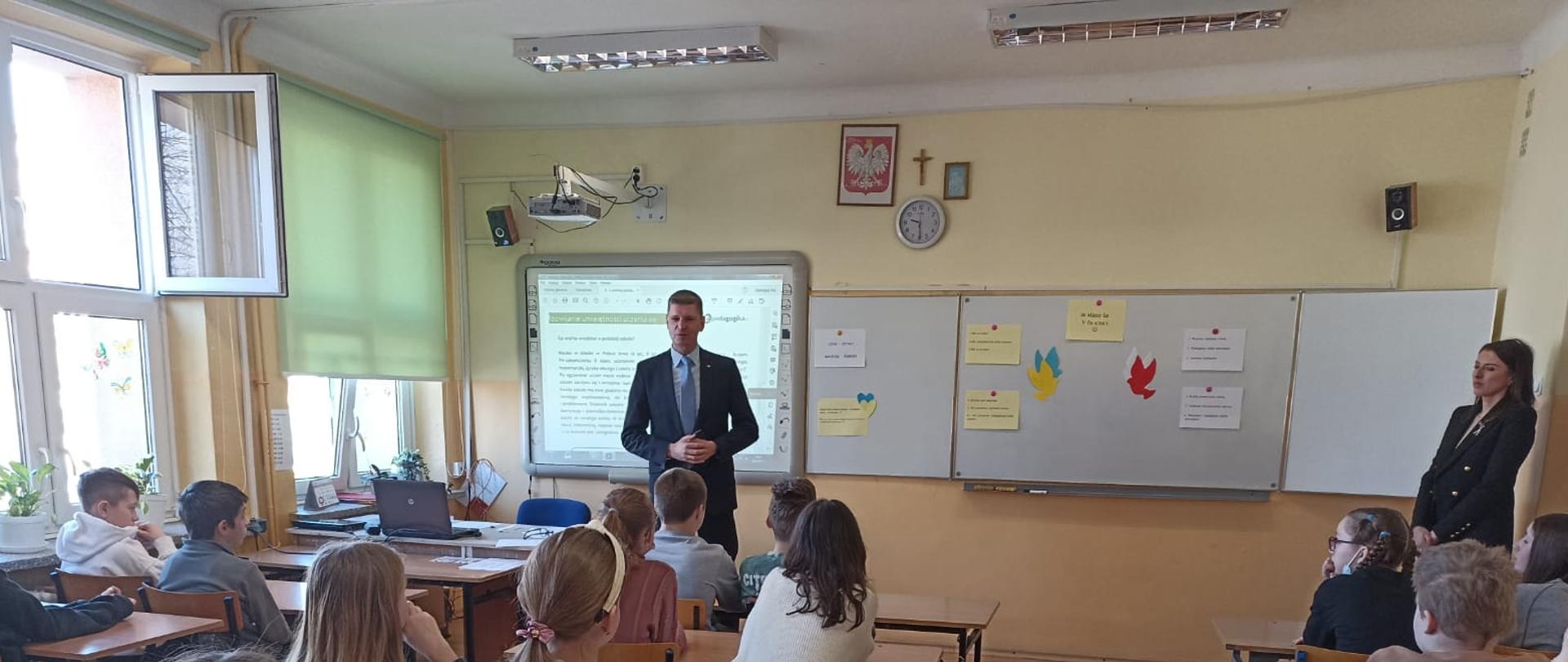 Klasa, grupa uczniów siedzących w ławkach. Przy tablicy multimedialnej stoi wicemimister Dariusz Piontkowski. Przy drzwiach nauczycielka w czarnych żakiecie. 