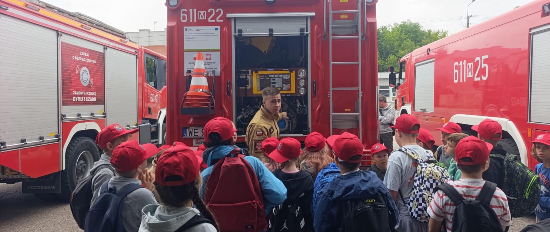 Zdjęcie przedstawia grupę dzieci uczestniczących w prelekcji edukacyjnej prowadzonej przez strażaka na placu manewrowym. W kadrze widoczne są trzy czerwone samochody ratowniczo-gaśnicze. W centrum stoi wóz o numerach operacyjnych 611[M]22 z otwartą tylną skrytką, w której znajduje się specjalistyczny sprzęt (żółty agregat). Strażak w piaskowym mundurze stoi przodem do dzieci i tyłem do pojazdu, opowiadając o swojej pracy. Grupa kilkunastu dzieci w wieku szkolnym, ubranych w sportowe stroje, plecaki oraz czerwone czapki z daszkiem, stoi półkolem i z zaciekawieniem słucha funkcjonariusza. Po lewej stronie widoczny jest fragment innego wozu strażackiego z plakatem promującym montaż czujek dymu i czadu, a po prawej stronie fragment trzeciego samochodu o numerach 611[M]25. Pogoda jest pochmurna.